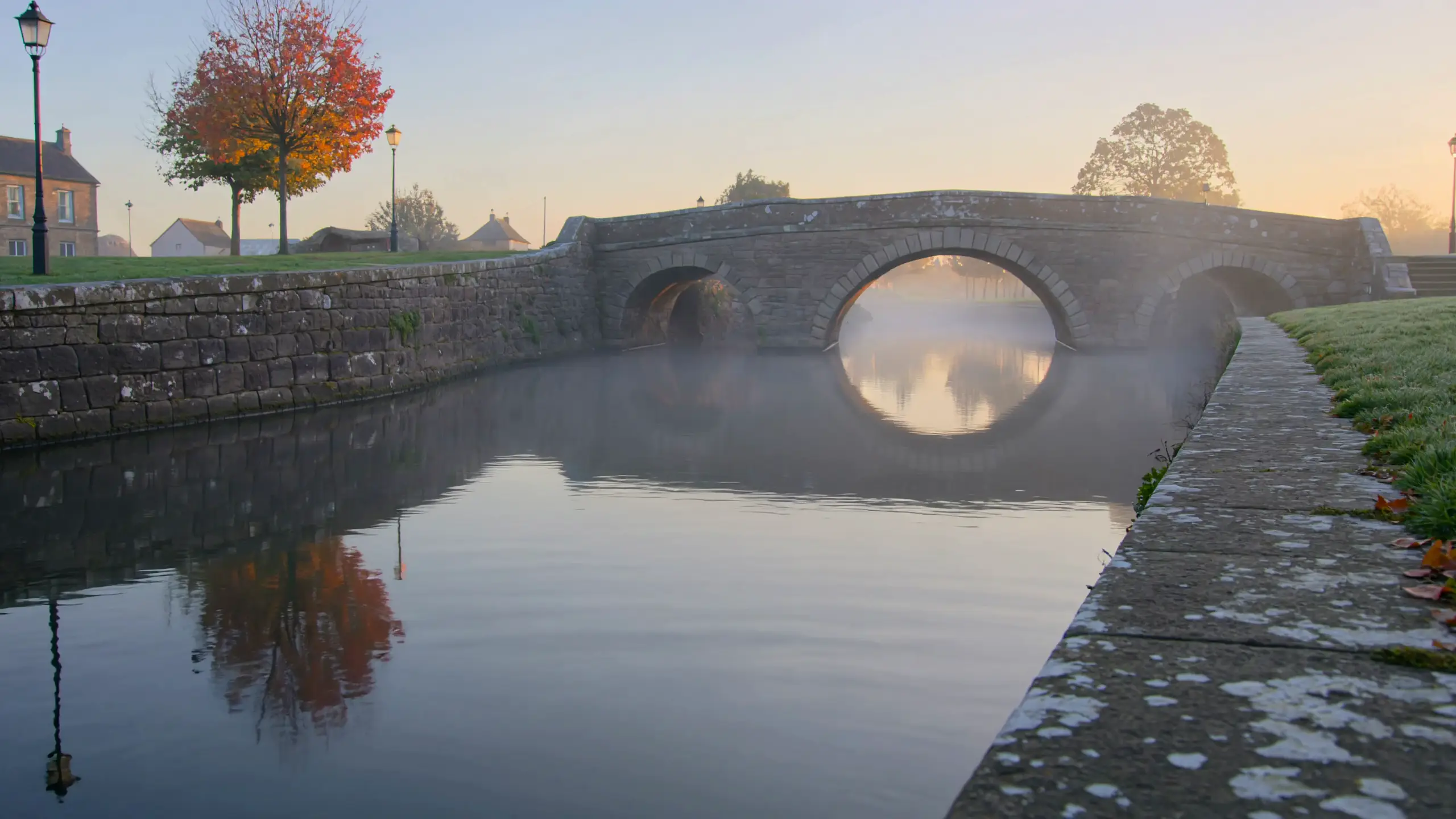 A stone bridge arches over a misty canal at sunrise, an autumn tree glowing red on the bank and the still water reflecting the sky.